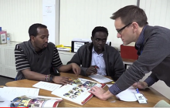 Adult ESL teacher talking to two learners sitting at a table