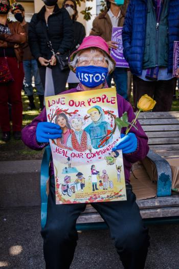 elderly person sitting on a bench holding a sign reading love our people heal our communities