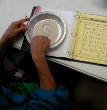 An adult student writes the letter S in sand with their finger