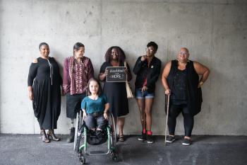 Six disabled people of color smile and pose in front of a concrete wall. Five people stand in the back, with the Black woman in the center holding up a chalkboard sign reading "disabled and here." A South Asian person in a wheelchair sits in front.