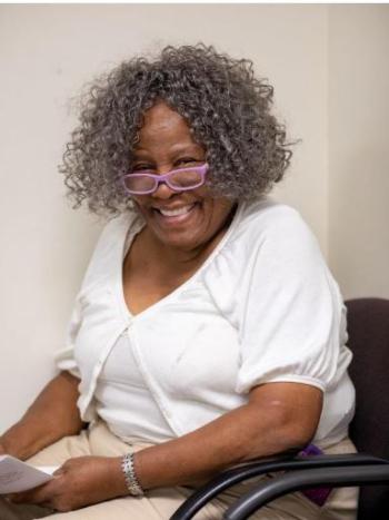 a smiling middle aged black woman with curly gray hair and purple glasses sits in a wheelchair and holds a piece of paper