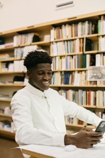 An adult student looks at his phone and smiles with a large shelf of books in the background. 