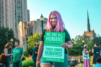 woman with pink hair holding a sign at a protest that reads we are all humans